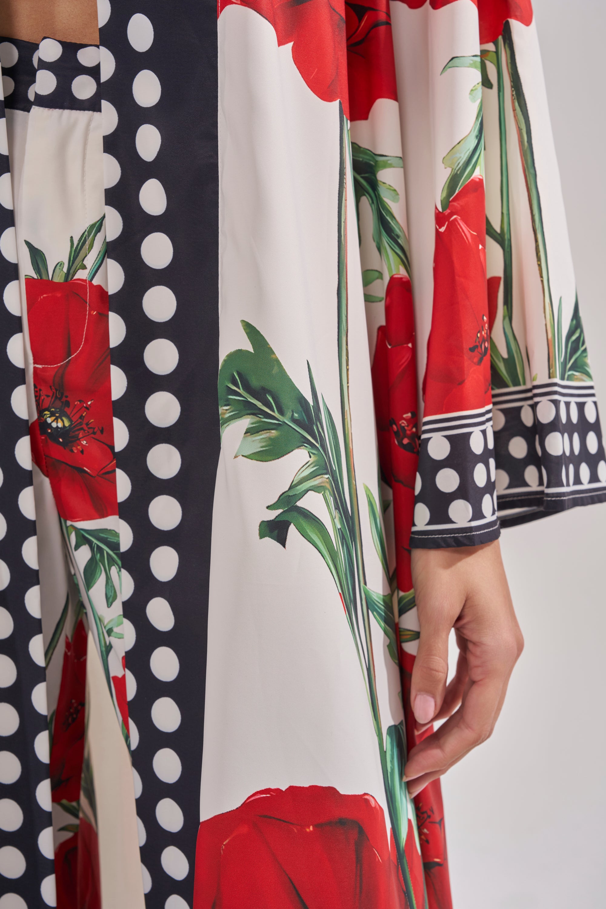 A close-up of a hand resting by the side, wearing the BELLADONNA CHIFFON DUSTER—a white vintage floral piece with bold red prints and black-and-white polka dot trim.