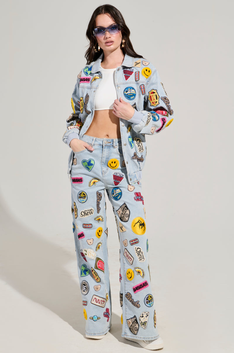 A woman wearing sunglasses, a white crop top, and the AUSTIN WIDE LEG PATCH DENIM jacket and pants set stands confidently against a plain background, showcasing her flair for high-rise waist fashion with colorful embroidered patches and logos.