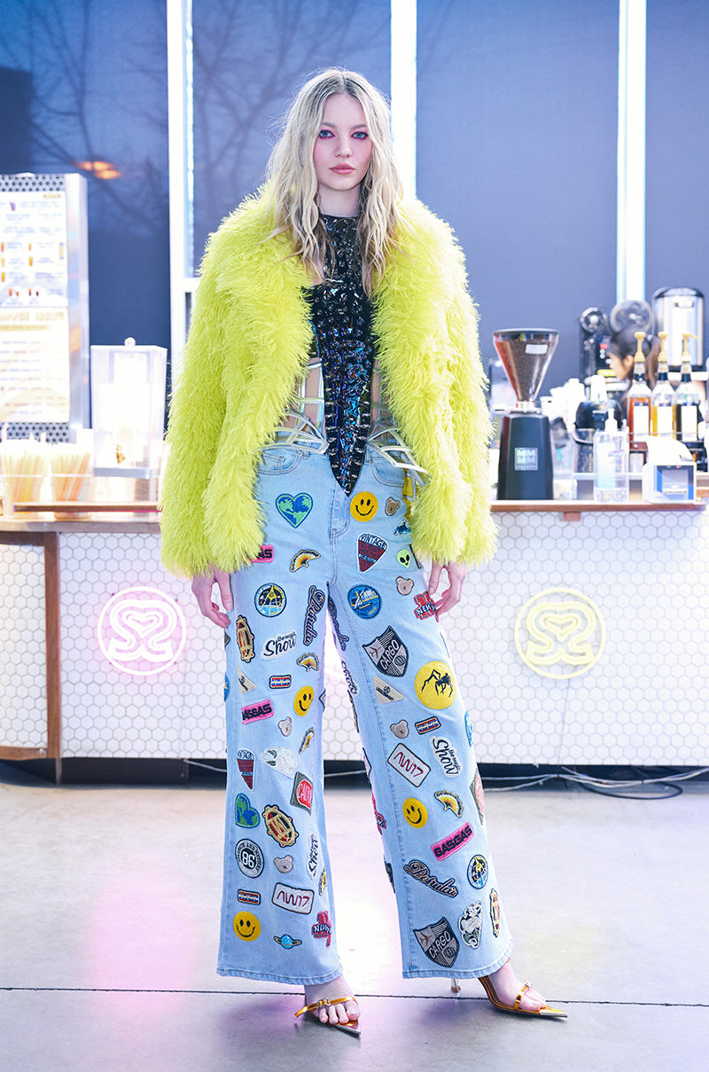 In a trendy, urban space with a counter featuring bottles and coffee equipment, an individual exudes confidence while sporting the AUSTIN WIDE LEG PATCH DENIM, characterized by its high-rise waist and vibrant embroidered patches. Their bright yellow furry jacket complements the modern setting perfectly.