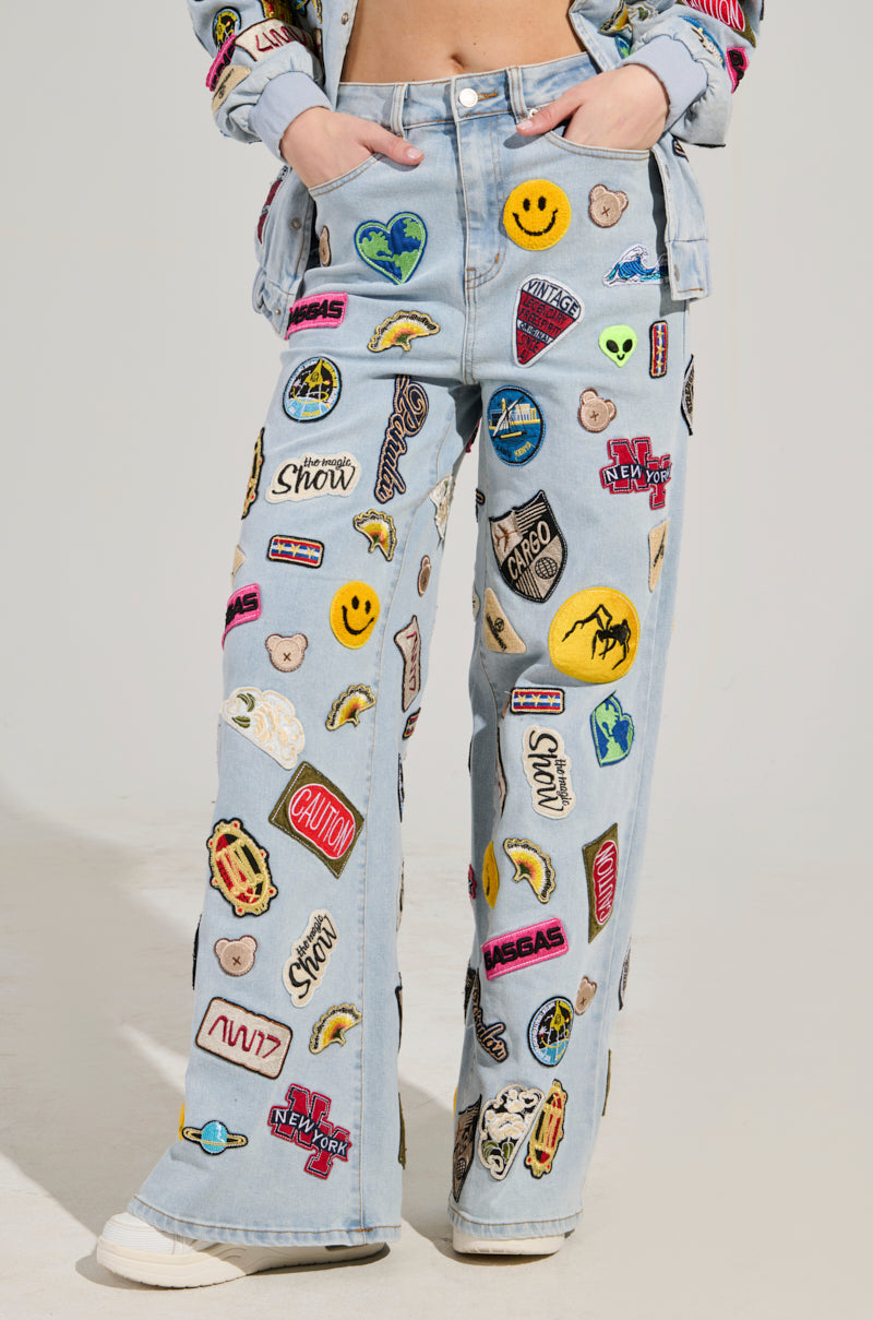 Someone is wearing the AUSTIN WIDE LEG PATCH DENIM, light blue jeans featuring high-rise waist and relaxed fit. These jeans are adorned with vibrant embroidered patches such as smiley faces, palm trees, and logos. They are ideally paired with a matching denim jacket flaunting similar lively embellishments.