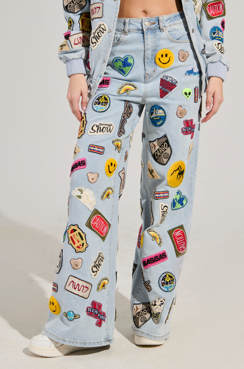 A person is wearing the AUSTIN WIDE LEG PATCH DENIM, featuring colorful embroidered patches such as smiley faces and logos. The high-rise waist complements a matching jacket that boasts similar vibrant patches. The plain background accentuates this lively ensemble.