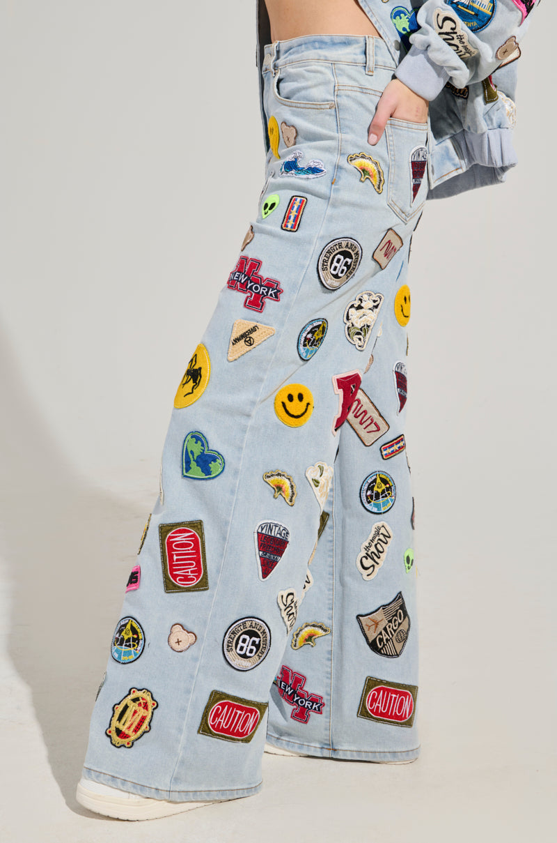 A person donning the AUSTIN WIDE LEG PATCH DENIM, featuring wide-leg jeans with a high-rise waist and vibrant patches such as smiley faces, caution signs, and various symbols. These jeans are perfectly paired with a matching denim jacket adorned with similar patches.