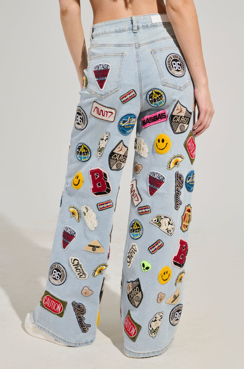 A person is standing against a plain background, wearing the AUSTIN WIDE LEG PATCH DENIM—light blue, high-rise waist, wide-leg jeans featuring a variety of colorful embroidered patches such as smiley faces, letters, and road signs.