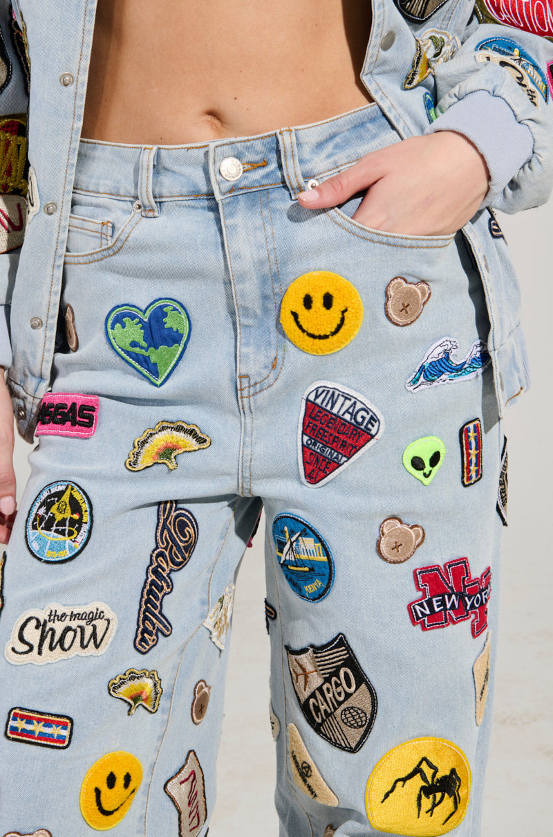 A person sporting the AUSTIN WIDE LEG PATCH DENIM, featuring a high-rise waist, paired with a jacket. Both pieces are embellished with vibrant embroidered patches including smiley faces, hearts, logos, and planet motifs. The individual's hand rests casually in a pocket.