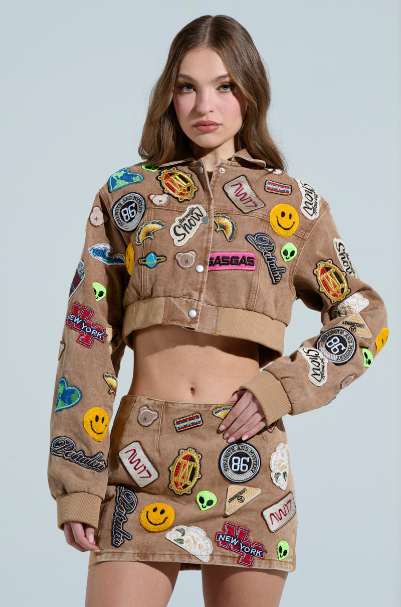 The woman wears an AKIRA Label ensemble—comprising the Meena Cropped Patch Denim Jacket and a matching skirt, both in brown with vibrant embroidered patches like smiley faces and logos. Standing confidently against a plain background, she looks forward with one hand on her hip.
