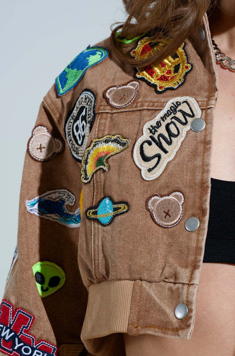 Wearing the MEENA CROPPED PATCH DENIM JACKET by AKIRA Label, featuring embroidered planets, a teddy bear, and "Shaw," paired with a black top and silver chain necklace creates an ensemble that showcases style and personality.