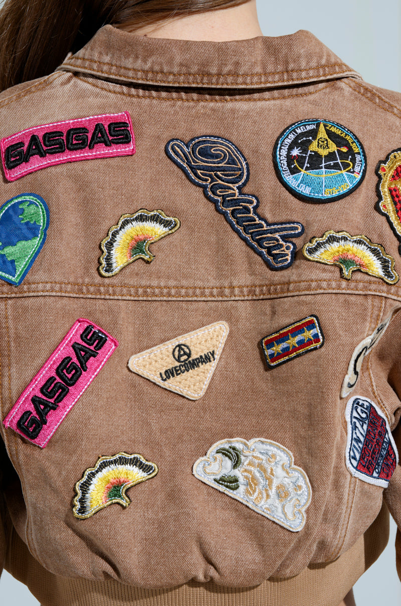 A person wearing the MEENA CROPPED PATCH DENIM JACKET, decorated with vibrant embroidered patches featuring text like "GASGAS" and "LOVEDOWNDAY," alongside planets and AKIRA Label patterns.