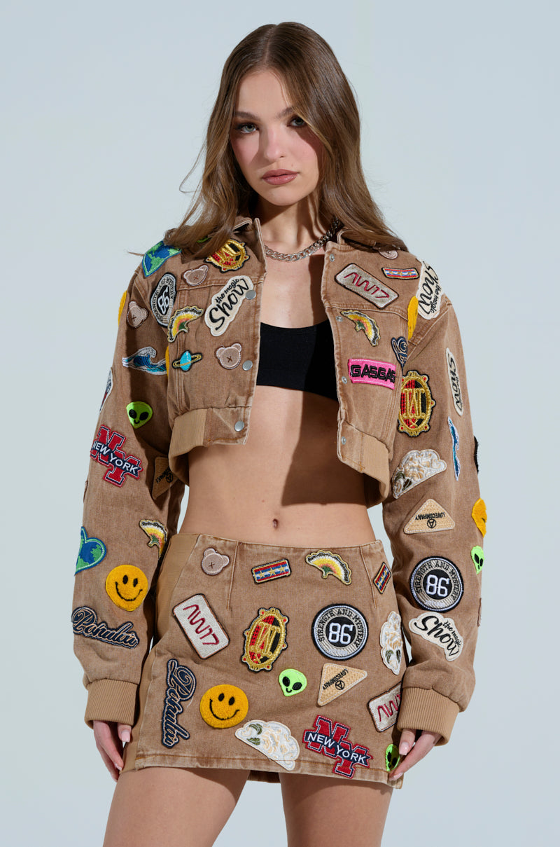 A woman in a brown outfit with vibrant embroidered patches stands against a plain background. She wears a black crop top beneath the MEENA CROPPED PATCH DENIM JACKET, showcasing designs like smiley faces and logos. Her expression is neutral, and her hair is loose.