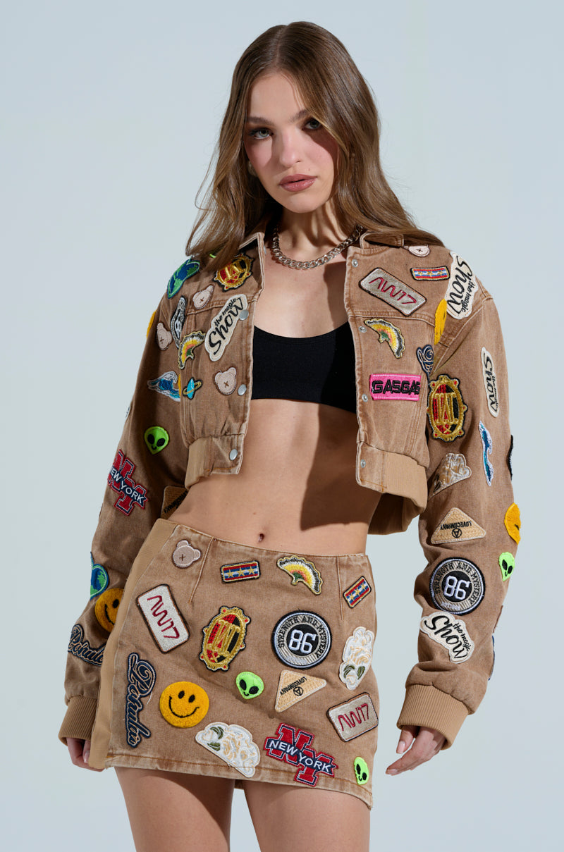 A woman poses against a light blue background, wearing the MEENA CROPPED PATCH DENIM JACKET and a matching skirt with vibrant embroidered patches. Her long hair flows over a black top, adding flair to her chic ensemble.