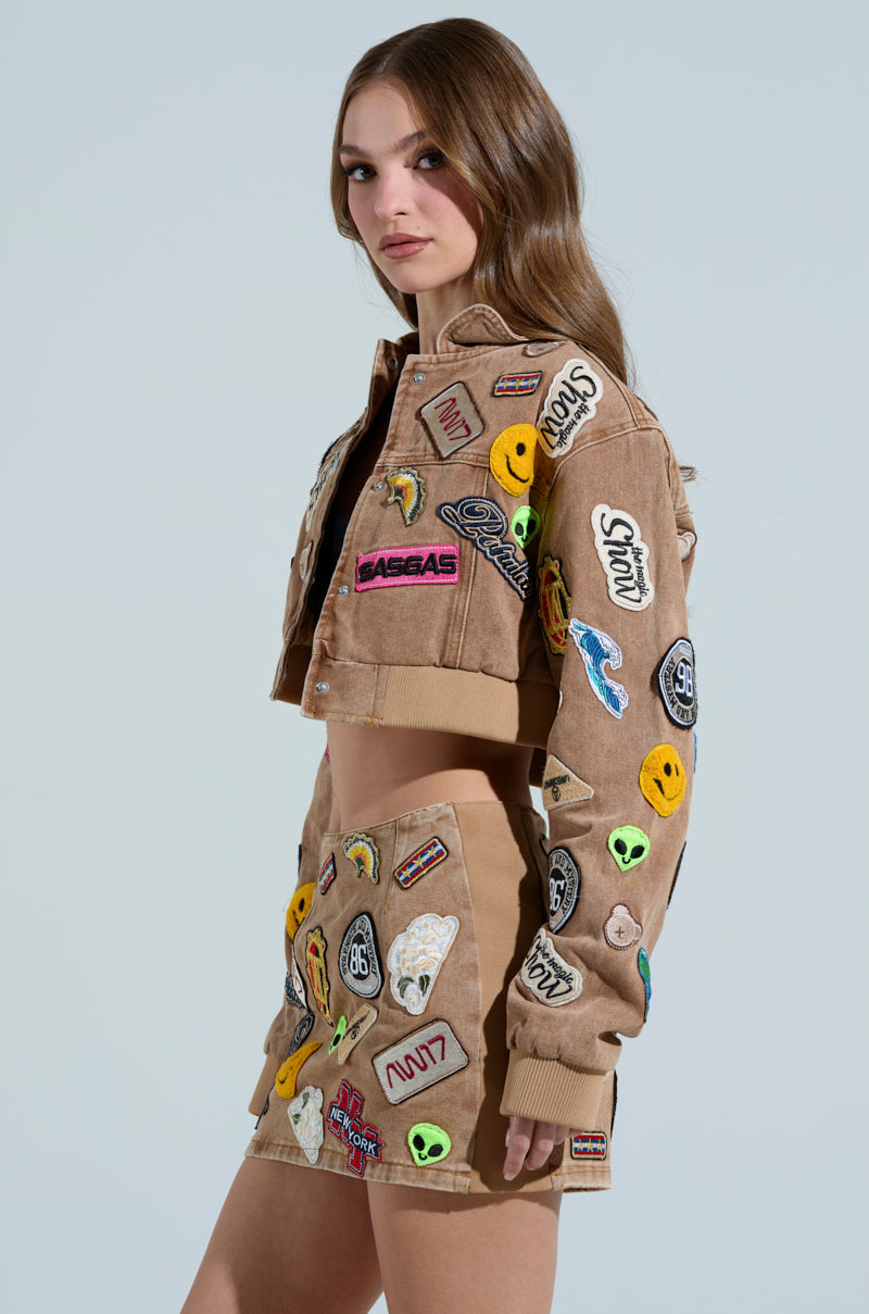 A woman with long brown hair models the MEENA CROPPED PATCH DENIM JACKET and a matching mini skirt, featuring embroidered patches of smiley faces, alien heads, and text, set against a light blue background.