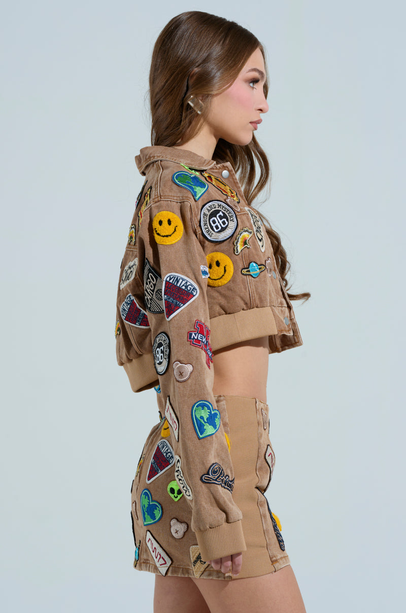 A woman in profile with long brown hair wears a MEENA CROPPED PATCH DENIM JACKET and matching shorts, adorned with colorful embroidered patches like smiley faces and logos, against a plain backdrop.