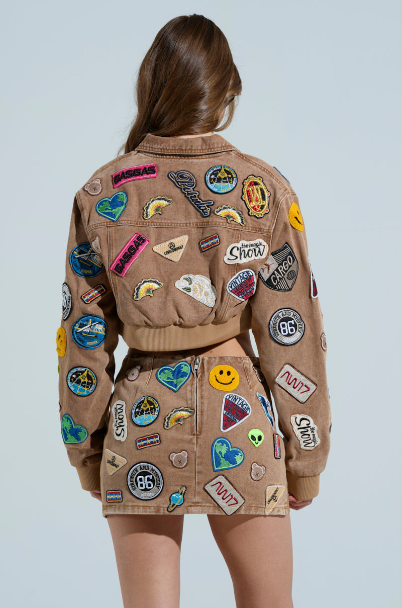 A woman with long brown hair, facing away, wears a MEENA Cropped Patch Denim Jacket and skirt adorned with embroidered patches like symbols, logos, and smiley faces, standing against a clear sky background.