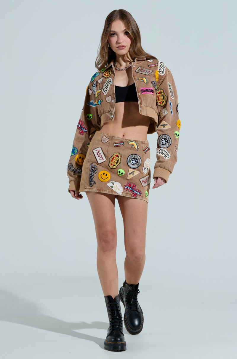 In a MEENA CROPPED PATCH DENIM JACKET, rich with vibrant embroidery reminiscent of the AKIRA Label style, paired with a black crop top and boots, a person confidently strolls against a plain, light-colored backdrop, embodying true urban chic.