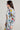 A woman confidently poses in the AUSTIN PATCH DENIM JACKET, a stylish piece of outerwear featuring light blue denim with various colorful embroidered patches and badges. Paired with matching pants, a white top, and sunglasses, she stands against a plain background.
