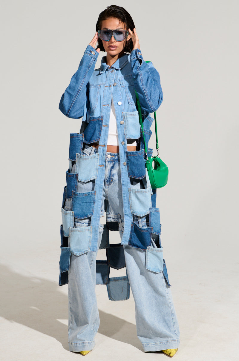 A person sporting the distinctive BRAXTON DENIM POCKET TRENCH, featuring a blend of various shades and patches, teamed with wide-leg jeans. They complete their look with sunglasses and a small green bag, posing stylishly against a plain background.