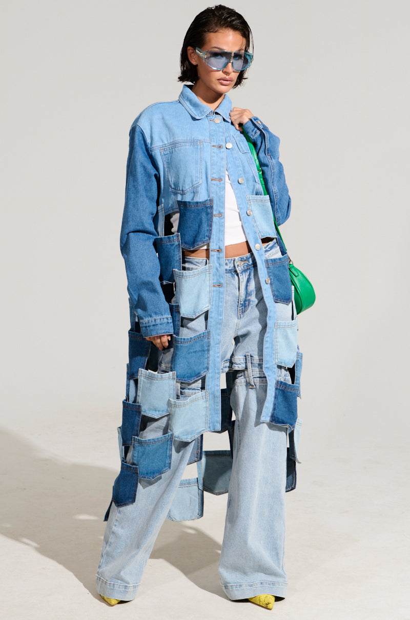 A person wearing a stylish AKIRA Label BRAXTON DENIM POCKET TRENCH and jeans, paired with a white top and blue sunglasses. They hold a green bag and wear yellow shoes. The plain background enhances the fashion ensemble.