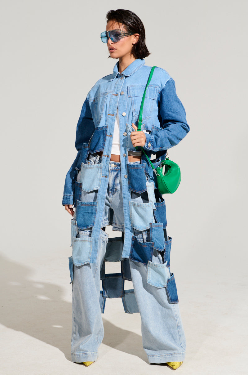 The person wears the BRAXTON DENIM POCKET TRENCH, a distinct denim trench coat with a mix of blue shades and large patchwork pockets. They complete their look with a green bag, blue-tinted sunglasses, and yellow shoes, all highlighting an AKIRA Label style against a simple backdrop.