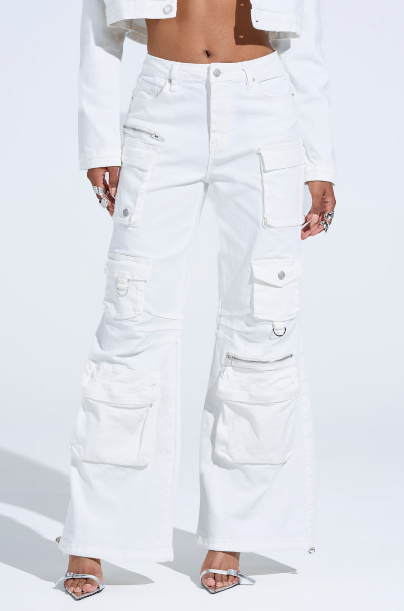 Someone effortlessly models KYLIE CARGO DENIM PANTS, stylishly paired with a matching crop jacket. These white cargo denim pants embody streetwear style with multiple pockets and zippers. Silver high-heeled sandals and various rings complete the look against a sunny, minimalist backdrop.