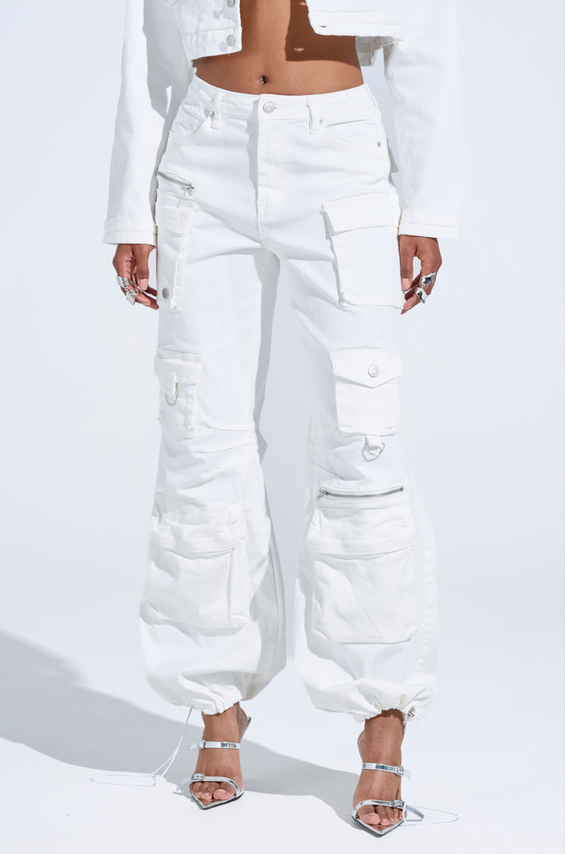 Someone in KYLIE CARGO DENIM PANTS with multiple pockets and zippers pairs them seamlessly with a white cropped jacket, completing the streetwear look with silver high-heeled sandals and silver rings against a plain background.