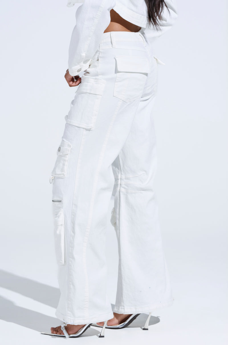 A person in the KYLIE CARGO DENIM PANTS and white high heels stands with one leg slightly crossed, showcasing a casual streetwear style against a plain backdrop. The white pants have multiple pockets and a relaxed fit.
