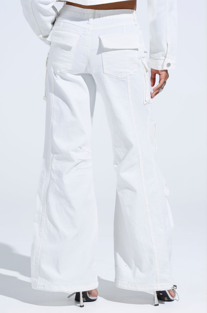 A back view of someone in KYLIE CARGO DENIM PANTS, high-rise wide-leg with multiple pockets, paired with a white jacket. They're confidently standing on a light surface, wearing stylish black and white heels.