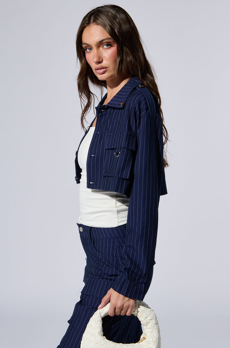 A woman with long dark hair is wearing the AKIRA Label RAYE PINSTRIPE CARGO BLAZER and matching pants over a white top. She is holding a small white woven handbag with metallic hardware and is looking slightly to her left. The background is plain gray.