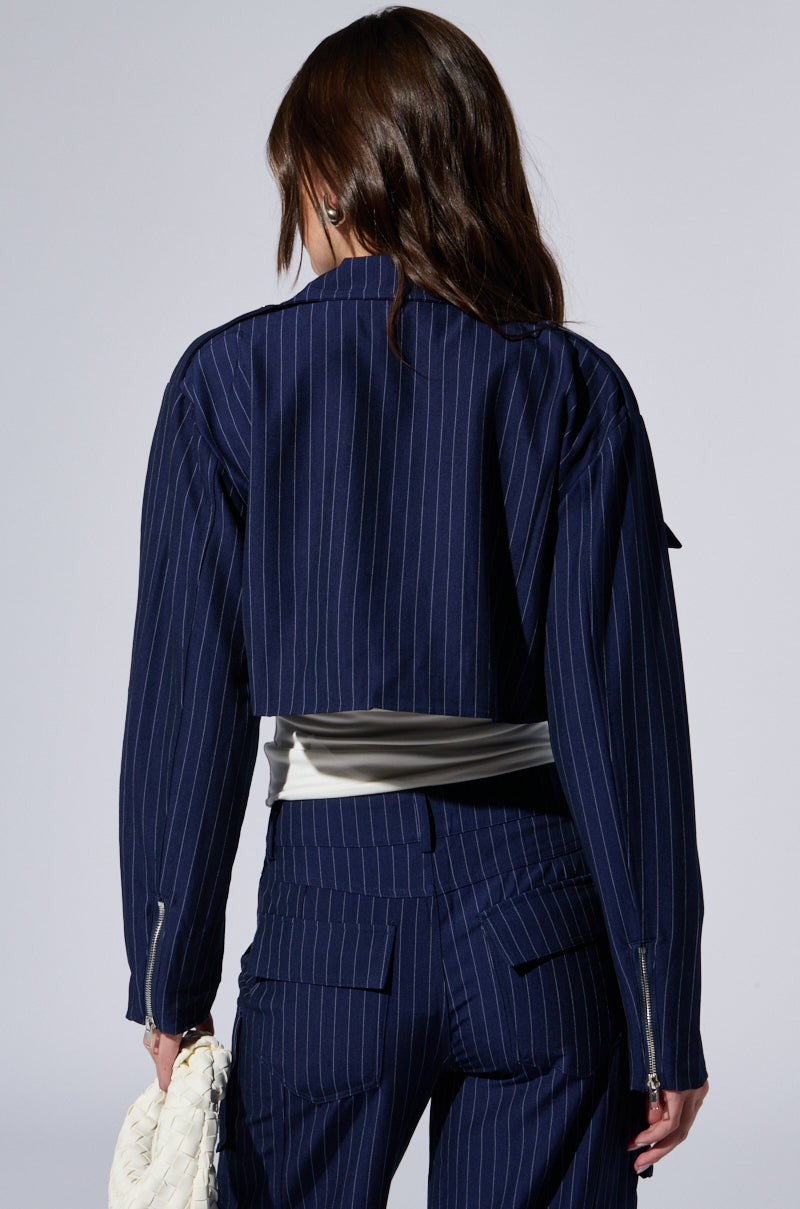 A person with long brown hair, seen from behind, wearing an AKIRA Raye Pinstripe Cargo Blazer. The cropped suit jacket is paired with matching pants and features metallic hardware. The individual holds a white textured bag in their left hand against a plain, light-colored background.