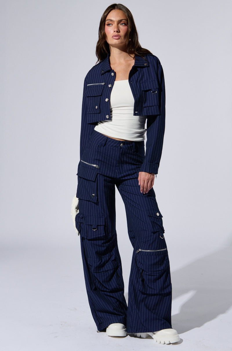 A woman stands confidently wearing the RAYE PINSTRIPE CARGO BLAZER and matching pants set from the AKIRA Label. She pairs the outfit with a white crop top and white sneakers. The jacket, adorned with pinstripe patterning and metallic hardware, features large front pockets, while the pants boast multiple pockets down the legs.