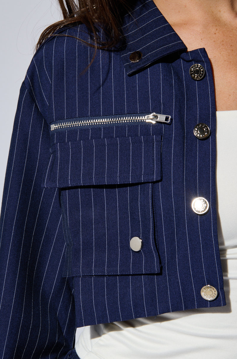 Close-up of someone wearing the RAYE Pinstripe Cargo Blazer from AKIRA Label, adorned with metallic hardware. The navy blue jacket showcases a flap pocket with a button closure and an additional zipper pocket above it. Underneath, they are dressed in a white top. Only part of the person is visible.
