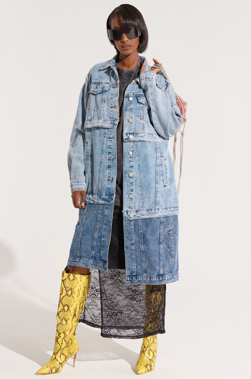Person wearing large dark sunglasses, a long, multi-tone denim trench coat named the "PULL UP LAYERED TRENCH" with functional pockets layered over a black dress with sheer lace at the bottom, and bright yellow snakeskin-patterned knee-high boots. Their right hand is resting on the trench coat's front, and the background is plain white.