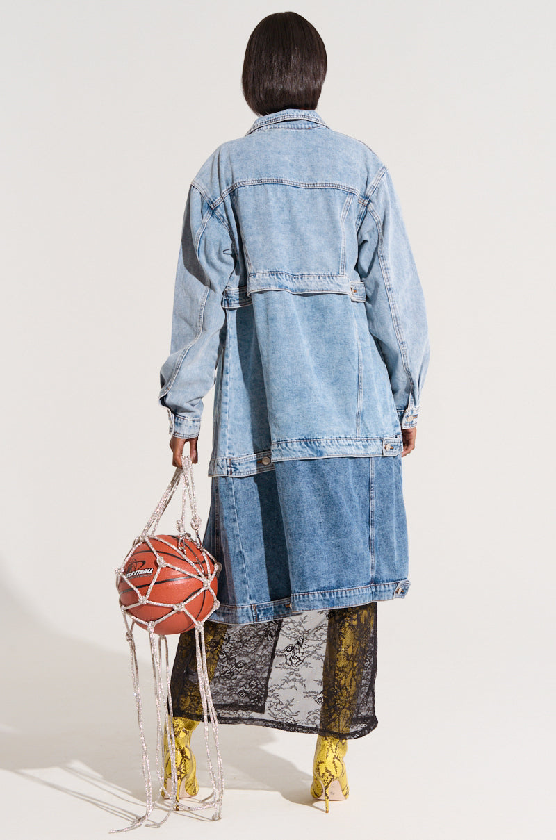 A person with short dark hair is seen from behind wearing a PULL UP LAYERED TRENCH, which is a long, light blue denim jacket featuring functional pockets and patches of darker denim. They complete their outfit with a black lace skirt and yellow snakeskin boots, while holding a handbag that resembles a deconstructed basketball with white netting straps.