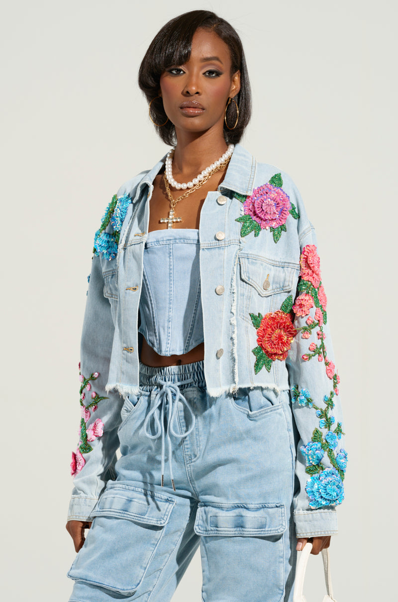 Wearing the GARDEN OF EDEN EMBELLISHED DENIM JACKET adorned with vibrant floral embroidery, they pair it with matching cropped silhouette pants. Complemented by a necklace and posing against a light background, they exude effortless style.