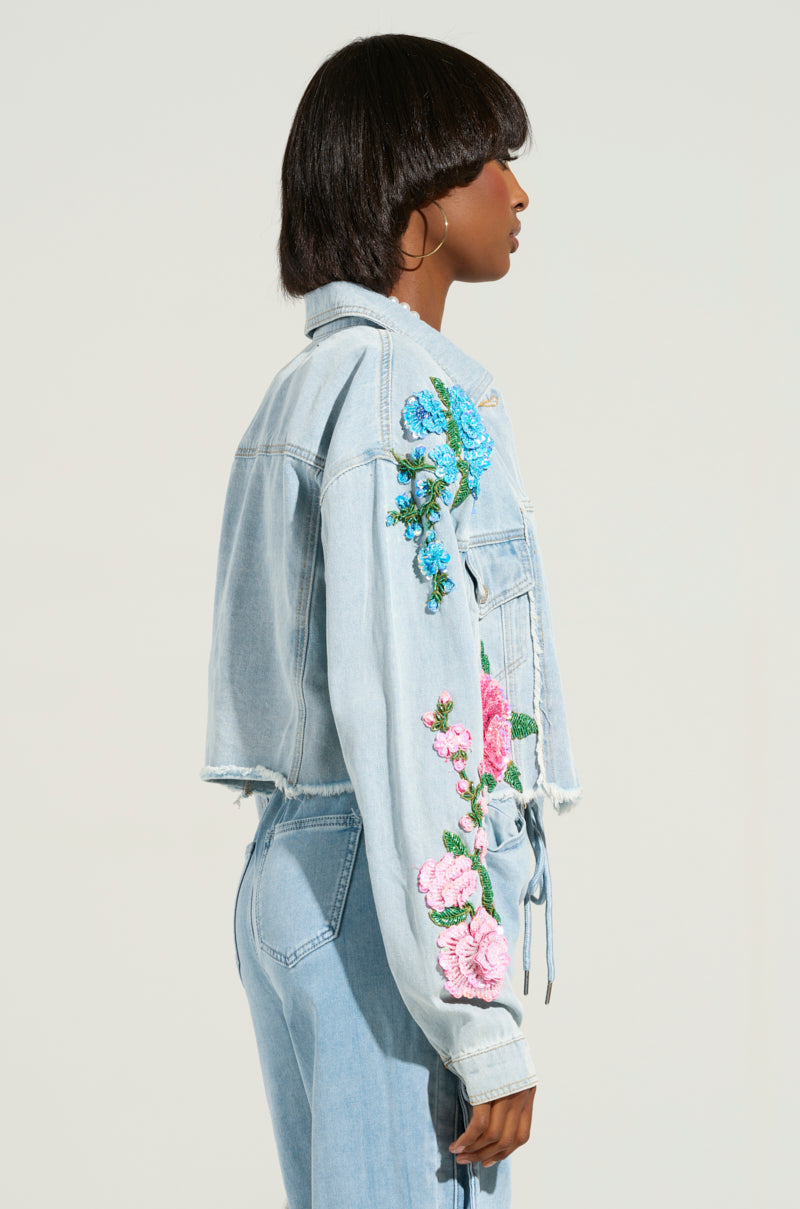 Against a neutral background, a person wears the GARDEN OF EDEN EMBELLISHED DENIM JACKET, featuring vibrant floral embroidery on the sleeves and shoulders. The cropped silhouette pairs seamlessly with their matching jeans, offering a stylish and cohesive look.