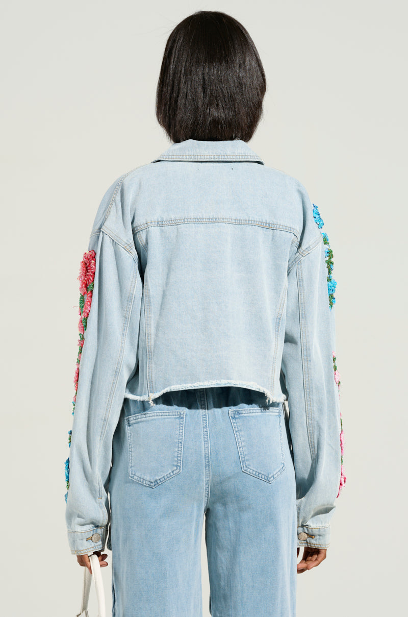 Someone with short dark hair is viewed from behind, highlighting the cropped silhouette of the GARDEN OF EDEN EMBELLISHED DENIM JACKET. This light blue denim jacket features vibrant floral embroidery on the sleeves and back, perfectly paired with matching denim jeans.