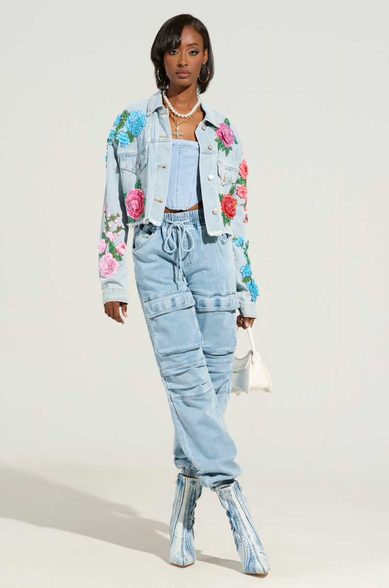 A woman stands confidently in the GARDEN OF EDEN EMBELLISHED DENIM JACKET, showcasing its chic, cropped silhouette with floral details. She carries a small white handbag and wears light blue boots that perfectly complement her short black hair and layered necklaces against a plain light background.