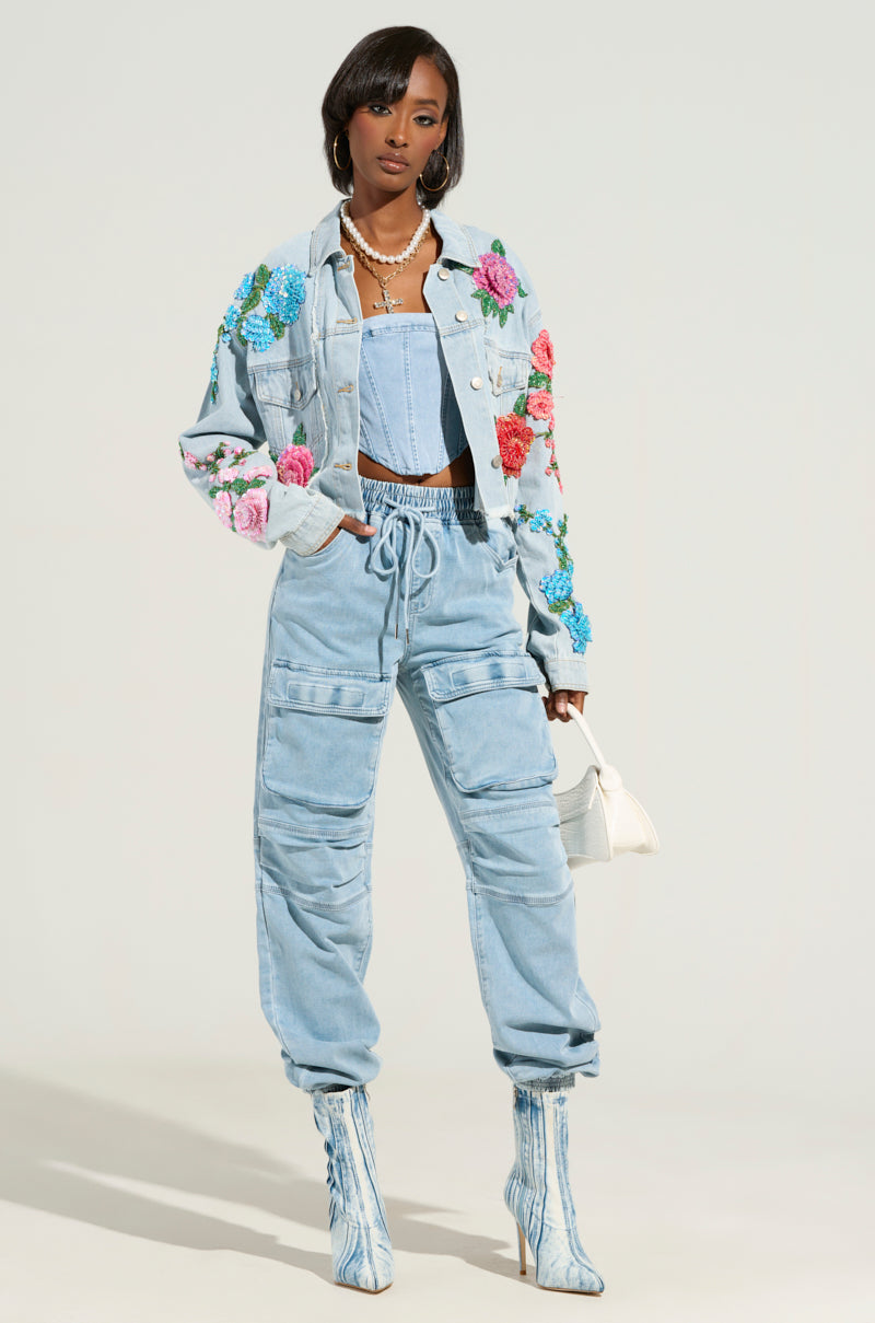 Wearing the Garden of Eden Embellished Denim Jacket, a person stands confidently in a light blue denim ensemble adorned with floral embroidery. They carry a small white handbag and wear matching denim boots, all set against a plain, neutral backdrop.