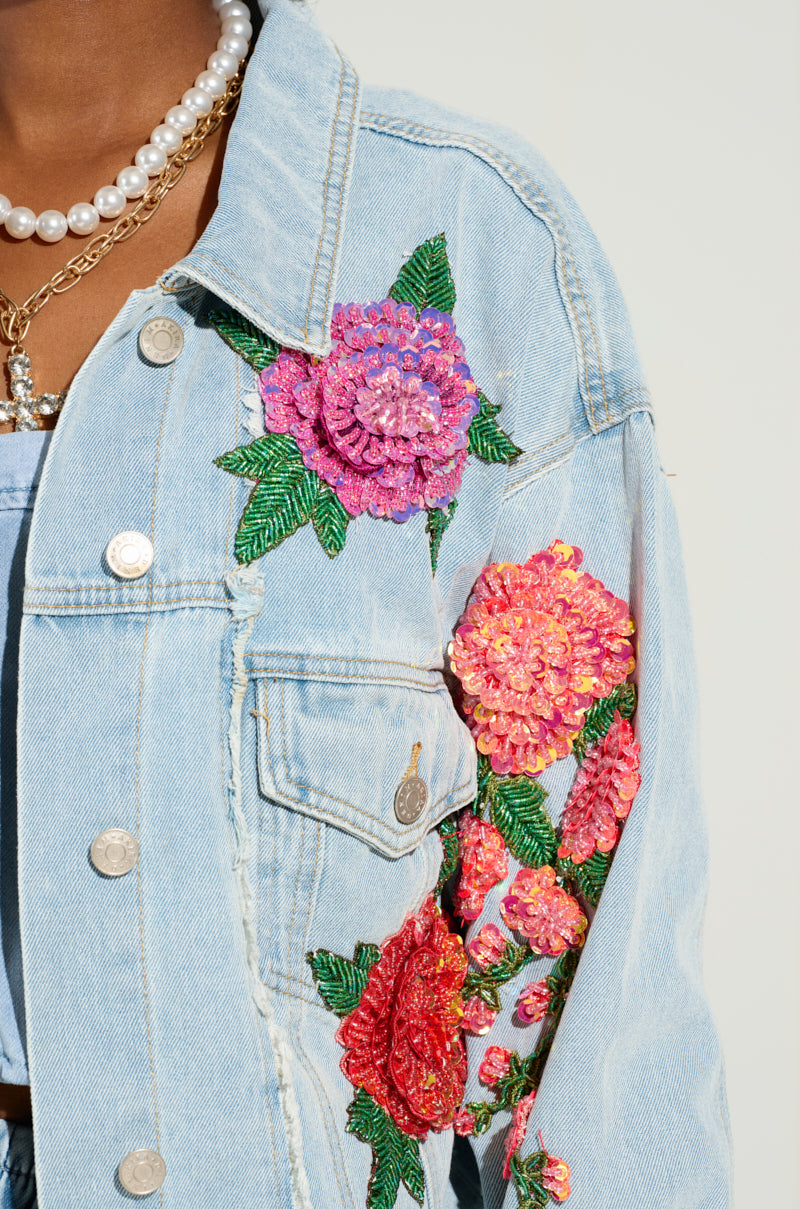 A person wears the GARDEN OF EDEN EMBELLISHED DENIM JACKET, a light blue denim piece adorned with colorful embroidered flowers and embellished floral designs. The jacket's silver buttons enhance its charm, and layered necklaces, featuring pearls and gold chains, add the finishing touch. Its cropped silhouette provides a modern twist.