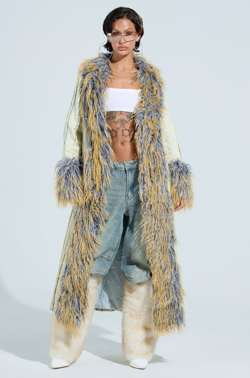 A person confidently poses in the GIA DISTRESSED DENIM TRENCH with a faux fur collar, paired with distressed denim jeans, a white bandeau top, sunglasses, furry leg coverings, and displaying chest tattoos.