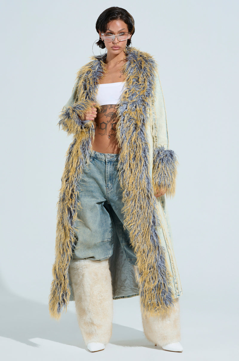 Confidently standing in a plain setting, a person showcases their bold style in hoop earrings, a white tube top, fluffy ankle-high boots, and the GIA DISTRESSED DENIM TRENCH topped with a long, colorful fur coat featuring a faux fur collar.