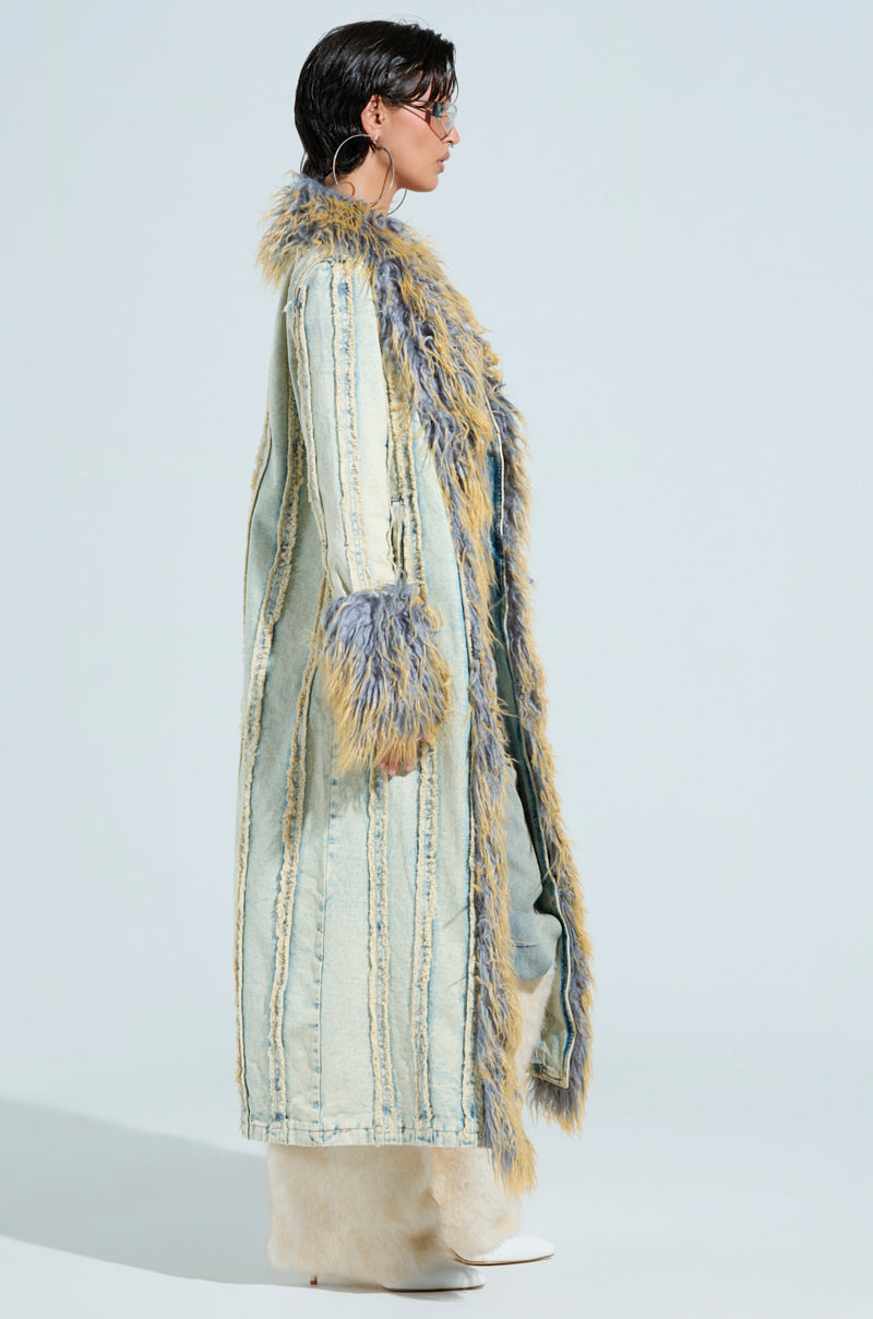 A person models the GIA DISTRESSED DENIM TRENCH featuring frayed edges and a faux fur collar. The coat's furry details also adorn the cuffs, complemented by white pants and shoes set against a neutral background.