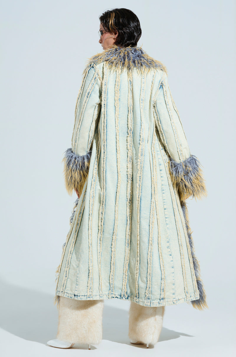 A person with long hair stands facing away, donning the GIA DISTRESSED DENIM TRENCH with a faux fur collar and cuffs. Vertical blue stripes adorn the coat, complementing their white fuzzy boots against a plain light grey background that highlights the stylish ensemble's visual appeal.