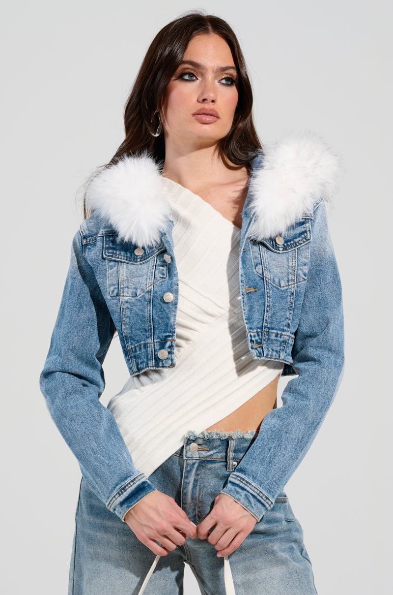 A person is wearing the SITTING PRETTY FUR TRIM DENIM JACKET over a white textured top and jeans, standing against a plain background. They gently hold the strings of the top, showcasing a stylish cropped silhouette.
