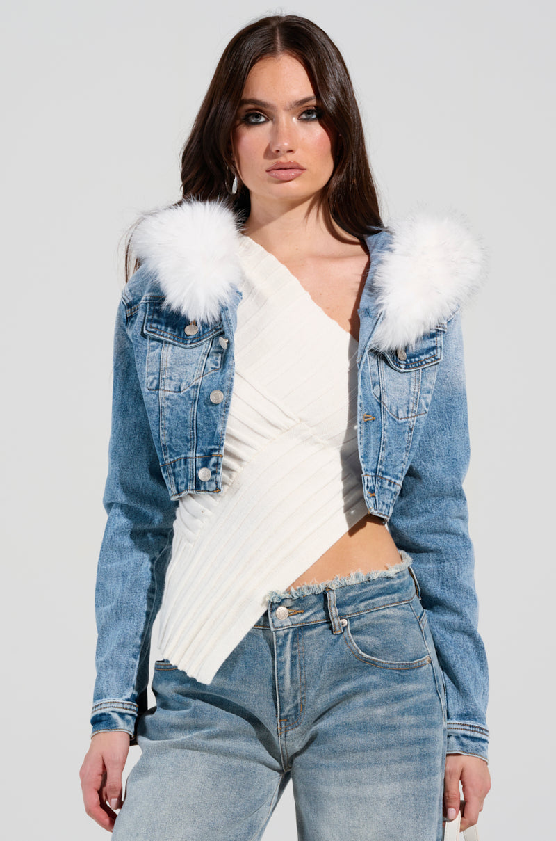 A person wearing the SITTING PRETTY FUR TRIM DENIM JACKET, featuring faux fur on the collar, styled with a white asymmetrical top and blue jeans against a plain light gray background.