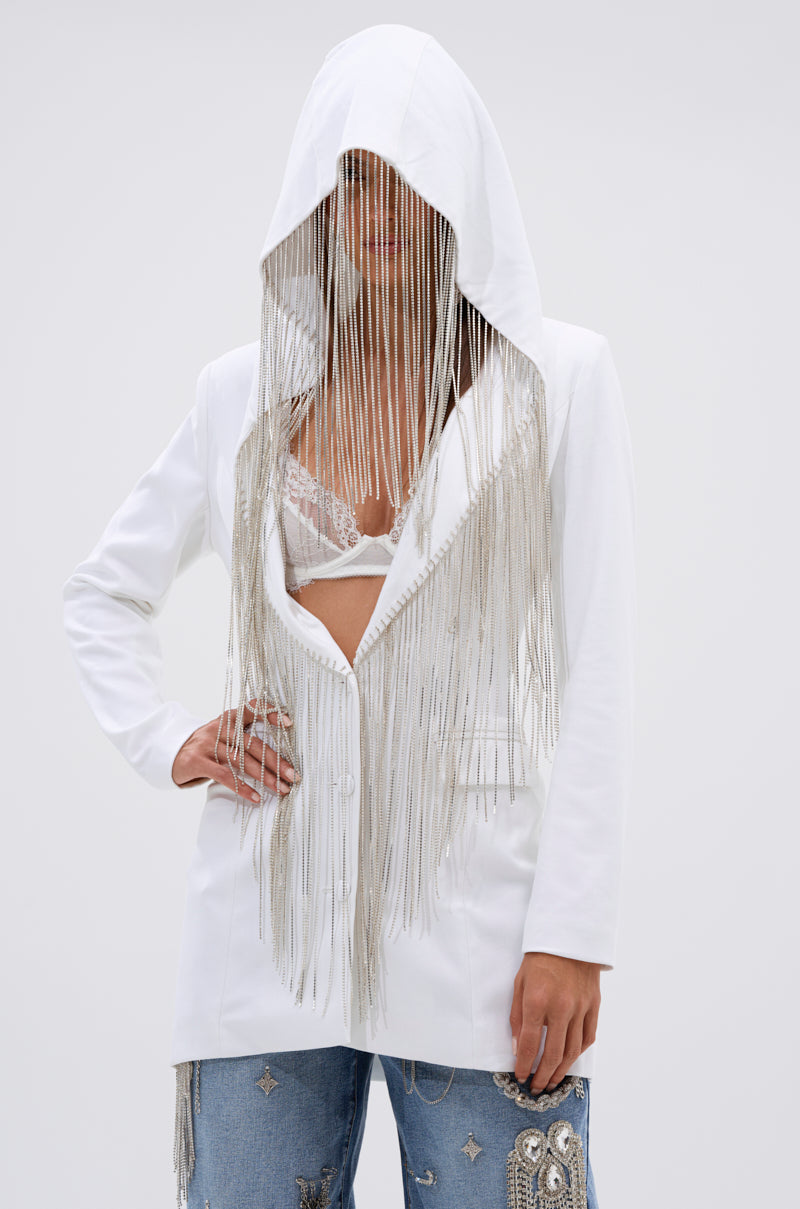 A person wears the DRIPPY RHINESTONE FRINGE BLAZER IN WHITE, with sparkling fringe on the hood over a lace bralette, paired with blue jeans decorated with embellishments. 1