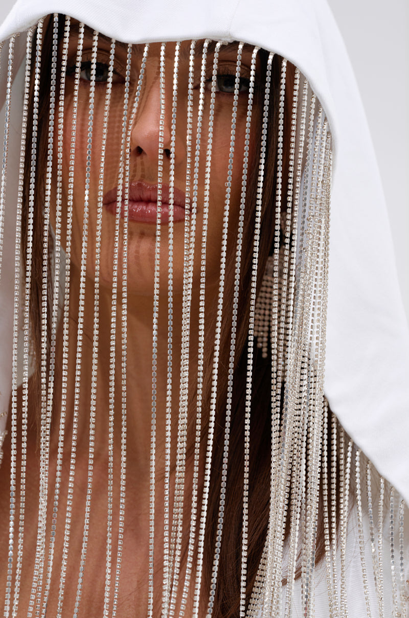 A woman models the DRIPPY RHINESTONE FRINGE BLAZER IN WHITE, featuring a hood and long sparkling fringe that partially covers her face. She has straight brown hair, neutral makeup, and closed eyes, set against a plain light background. 9