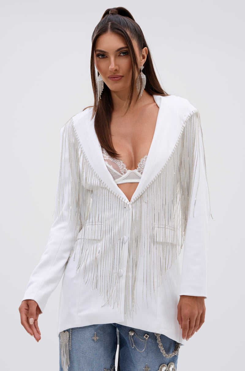 A woman poses confidently against a plain light gray background wearing the DRIPPY RHINESTONE FRINGE BLAZER IN WHITE, paired with a white lace bralette and blue jeans adorned with silver embellishments. 2
