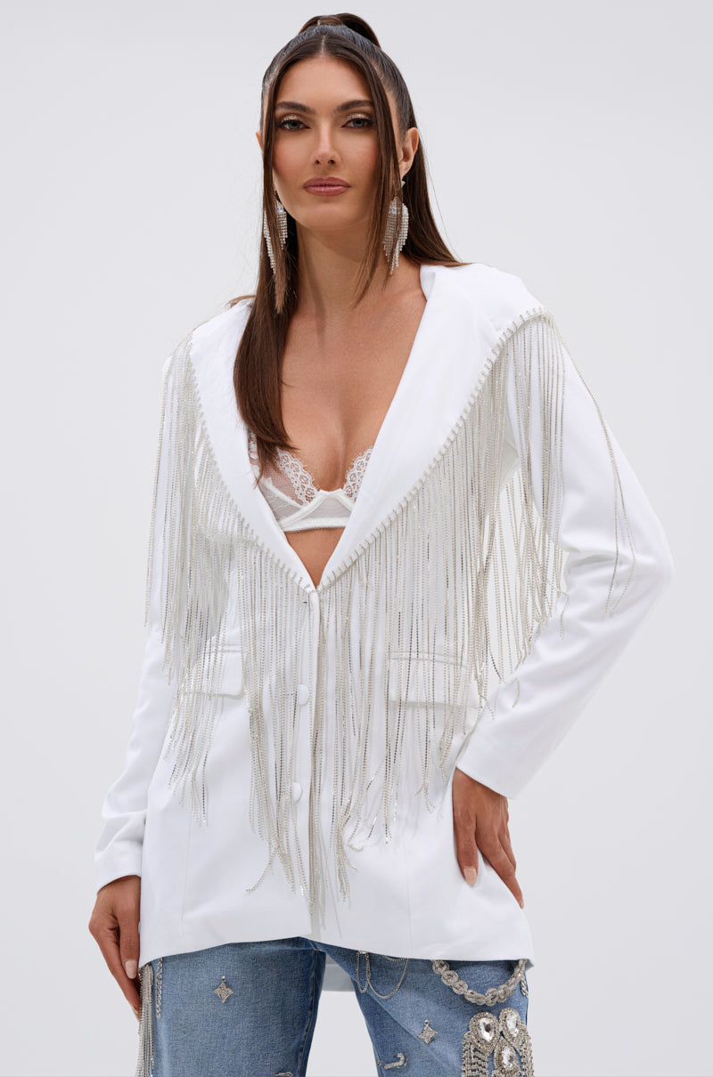 A woman poses confidently in the DRIPPY RHINESTONE FRINGE BLAZER IN WHITE with long fringe, paired with a white lace bralette and rhinestone-embellished blue jeans against a plain background. 0