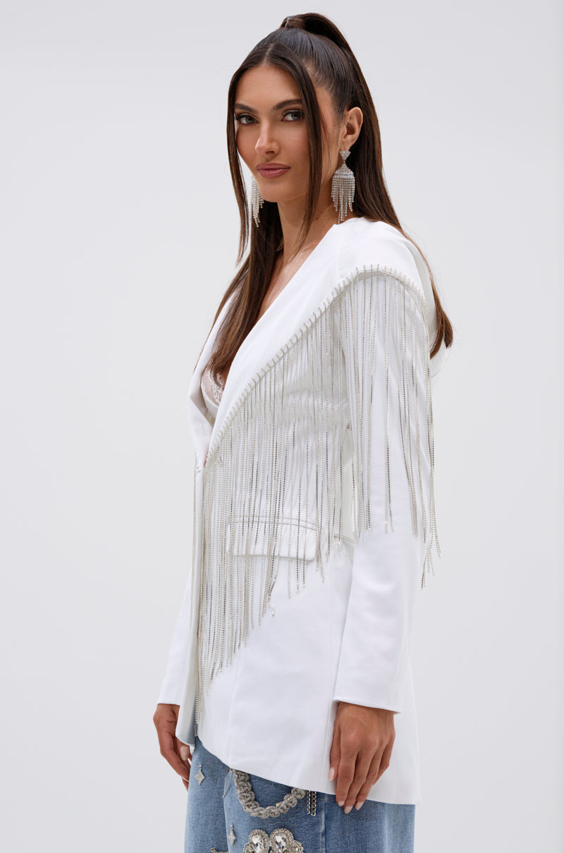 A woman with long brown hair in a high ponytail models the DRIPPY RHINESTONE FRINGE BLAZER IN WHITE, paired with statement earrings and embroidered blue jeans, against a plain light background. 4