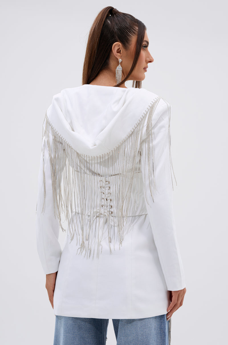 A woman with long brown hair in a high ponytail models the DRIPPY RHINESTONE FRINGE BLAZER IN WHITE, paired with dangling earrings and light blue jeans, facing away from the camera. 5