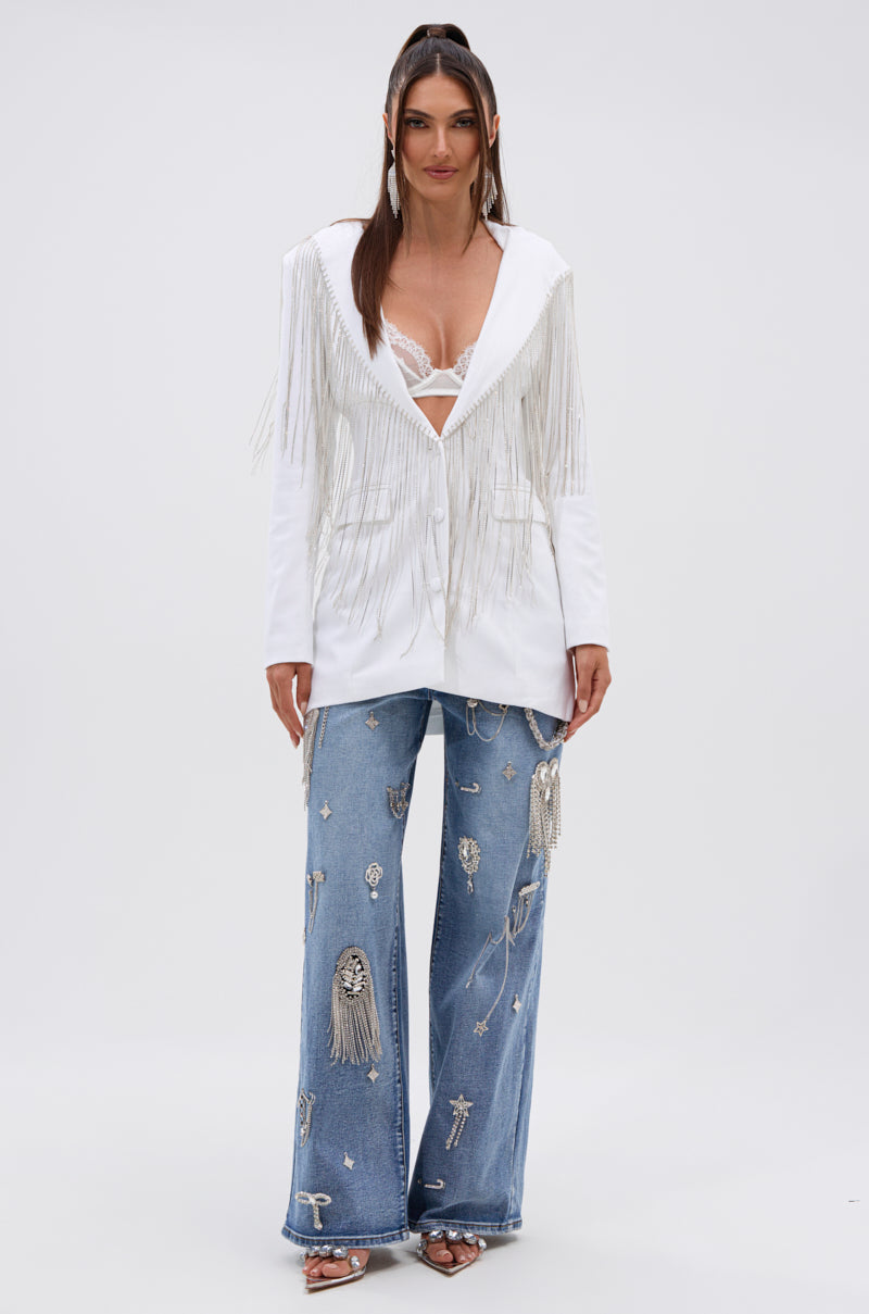 A woman with long brown hair wears the DRIPPY RHINESTONE FRINGE BLAZER IN WHITE over a visible white bra, paired with embellished light blue wide-leg jeans and heeled sandals, standing facing forward. 6